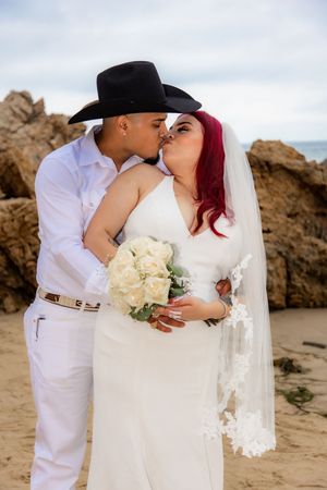 Bride and groom kissing on the beach, she holds white roses bouquet wearing veil, he wears black cowboy hat.