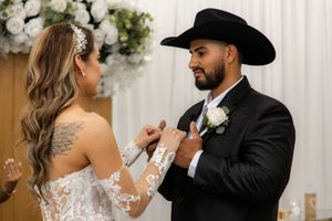 Groom in black cowboy hat smiling as bride adjusts his boutonniere during wedding ceremony with white flowers.