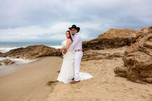 Wedding couple embracing on sandy beach with rocky coastline, groom in white outfit with black cowboy hat.