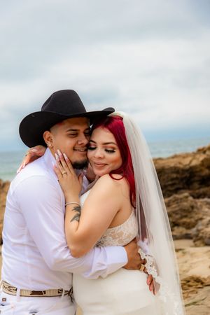 Bride with red hair and veil tenderly embraces groom in black cowboy hat on a rocky beach setting.