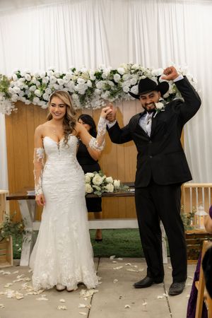 Newlyweds celebrating joyfully at altar with groom raising fist in triumph beneath white floral wedding arch.