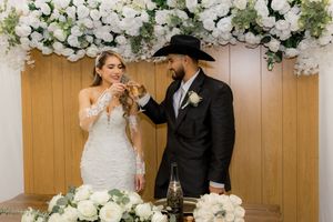 Bride and groom toasting with glasses at wedding ceremony table decorated with elegant white rose arrangements.