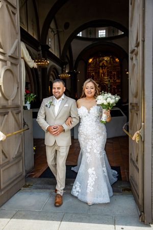 Smiling bride and groom exit a historic church doorway, bride holding white floral bouquet.