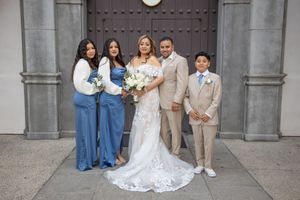Bride with wedding party including two bridesmaids in blue, groom, and ring bearer outside church.
