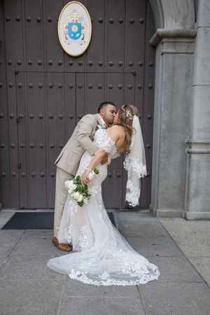 Groom dips bride in a romantic kiss outside ornate church doors after wedding ceremony.