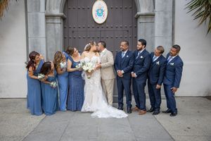 Wedding party in blue bridesmaids dresses and navy suits kissing outside a historic church.