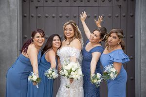 Bride poses joyfully with four bridesmaids in blue dresses in front of large wooden church doors.