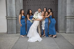 Groom surrounded by bride and four bridesmaids in blue gowns embracing outside a stone church.