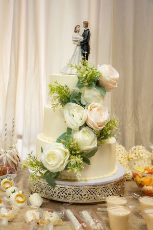 Elegant three-tier white wedding cake with roses and bride and groom topper on decorative gold stand.