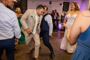 Guests dancing energetically at wedding reception with bride visible in white gown in background.