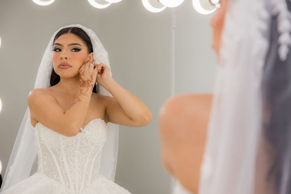 Bride in a strapless lace corset gown adjusts her earring while looking into a lighted vanity mirror.