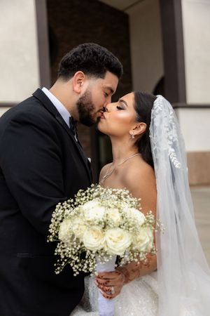 Bride and groom share a romantic kiss, bride holding white rose and baby's breath bouquet in elegant wedding attire.