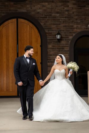 Bride and groom walk hand in hand before arched wooden doors, bride in sparkling ballgown with veil.
