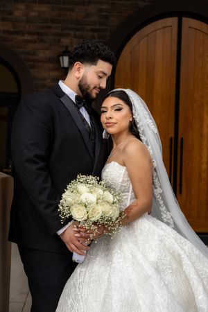 Elegant couple pose together by wooden doors, bride in lace ballgown holding white roses and baby's breath.