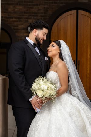 Bride and groom share a loving gaze beside arched wooden doors, bride in strapless lace ballgown with veil.