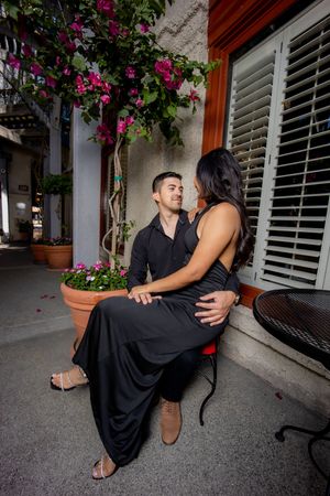 Couple dressed in black sitting closely together on outdoor patio surrounded by colorful bougainvillea flowers.