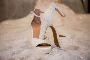 Elegant white studded bridal heels with ankle strap displayed on wedding dress fabric.