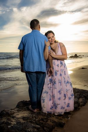 Woman in floral pink dress leans on partner's shoulder during romantic beach sunset photo session.