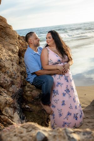 Couple shares a tender moment leaning against coastal rocks with ocean waves and sunset in background.
