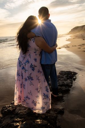Couple embraces in romantic silhouette against a stunning golden sunset on a scenic rocky beach.
