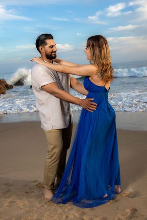 Couple dancing on beach at sunset, woman in flowing blue dress, man in beige shirt and khaki pants.