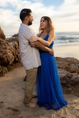 Laughing couple embracing near ocean rocks, woman in elegant blue tulle gown at golden hour beach.