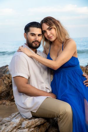 Romantic couple posing on beach rocks, woman in blue dress rests head on seated mans shoulder.