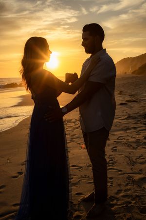 Silhouette of couple holding hands facing each other on beach during stunning golden sunset.
