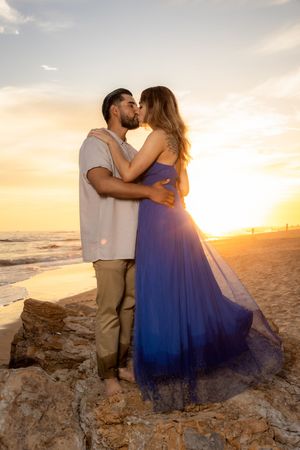 Couple sharing a kiss on rocky beach at sunset, woman in flowing blue dress, warm golden light.