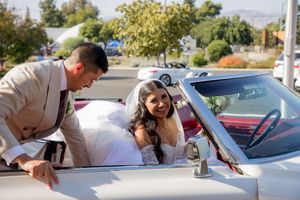 Bride in white wedding gown sitting in a classic convertible car while groom leans in smiling.