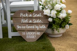 Heart-shaped wooden wedding sign reads Pick a Seat Not a Side beside white floral arrangement on grass aisle.
