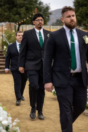 Three groomsmen in black suits with green ties walk down sandy outdoor wedding aisle lined with white flowers.