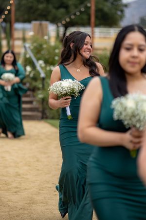 Bridesmaids in emerald green gowns carry white baby's breath bouquets while walking down sandy outdoor aisle.