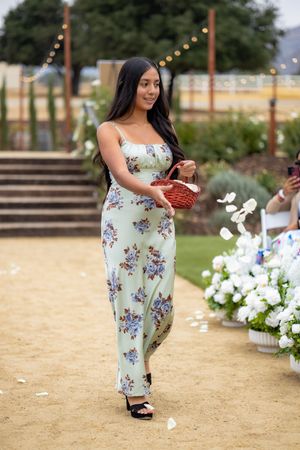 Young woman in floral mint maxi dress carries wicker basket as flower girl down sandy outdoor wedding aisle.