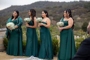 Four bridesmaids in emerald green gowns hold white bouquets standing at outdoor hilltop wedding ceremony.