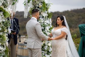 Bride and groom exchange vows under a white floral arch at an outdoor vineyard wedding ceremony.