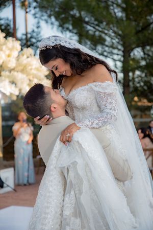Groom lifts bride in stunning lace wedding gown during their first dance at outdoor reception.