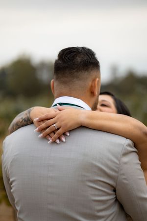 Bride embraces groom from behind showing diamond engagement ring at an outdoor wedding celebration.