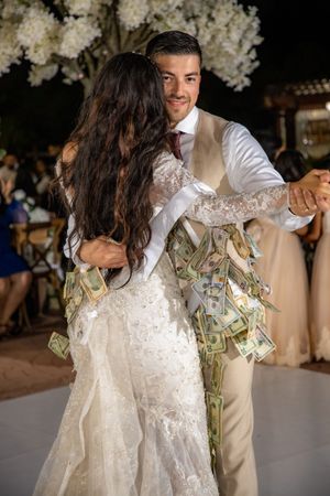 Bride and groom share first dance as guests pin money on wedding dress at reception.