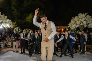 Groom in tan suit performs energetic dance move on white dance floor at wedding reception.