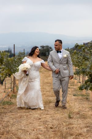 Bride and groom walk hand in hand through vineyard rows laughing on their wedding day.