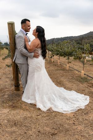 Bride and groom share a romantic moment leaning against a wooden post in a vineyard.