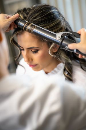 Stylist curls dark highlighted hair of bride during elegant wedding morning preparation session.