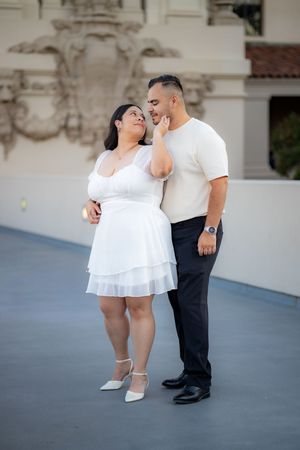 A couple poses romantically outdoors, woman in white mini dress and man in cream shirt with black pants.