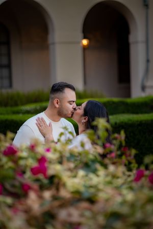 Couple shares a romantic kiss in a garden courtyard surrounded by colorful blooming flowers.