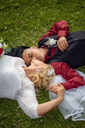 Newlyweds lie on green grass holding hands, groom in red shirt with boutonniere, bride in lace.