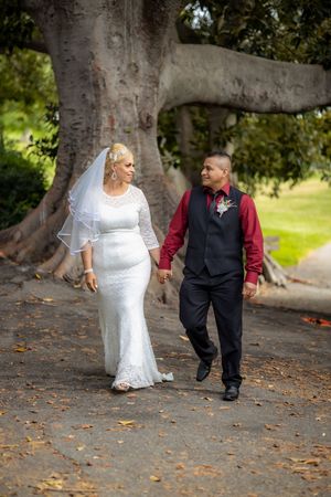 Bride in white lace dress and groom in burgundy shirt walk hand in hand under large tree.