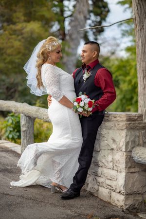 Bride in white lace gown and groom in red shirt pose together on a stone bridge in a garden.