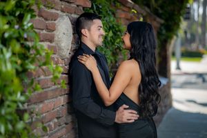 Couple in black outfits embracing against a brick wall with green ivy, sharing a romantic moment outdoors.