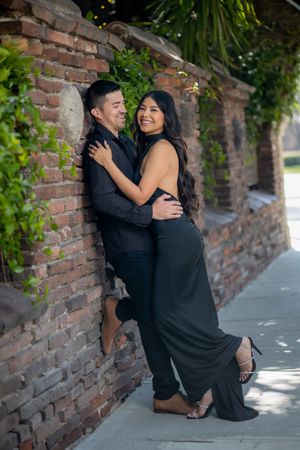Smiling couple in matching black formal wear posing together against a vine-covered brick wall outdoors.